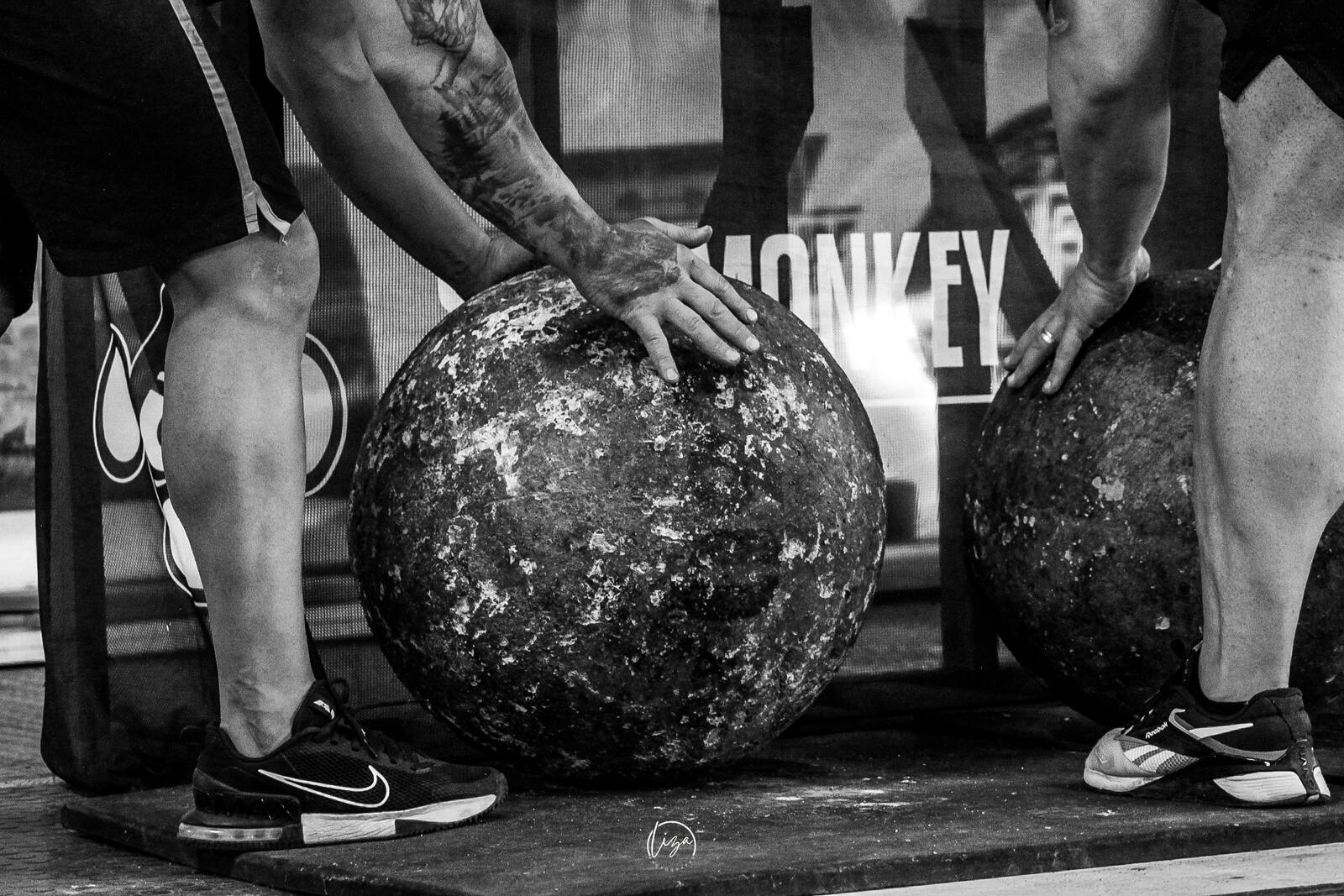 A chalked, tattooed hand resting on a weathered Atlas stone between an athlete's braced legs.