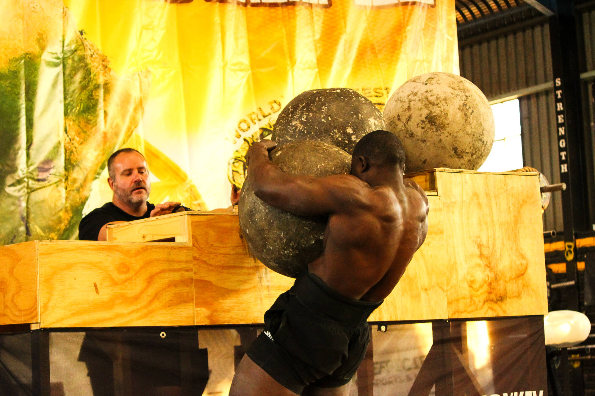 A strongman hoisting a loaded Atlas stone to his chest, mid-lift against a blazing arena backdrop.